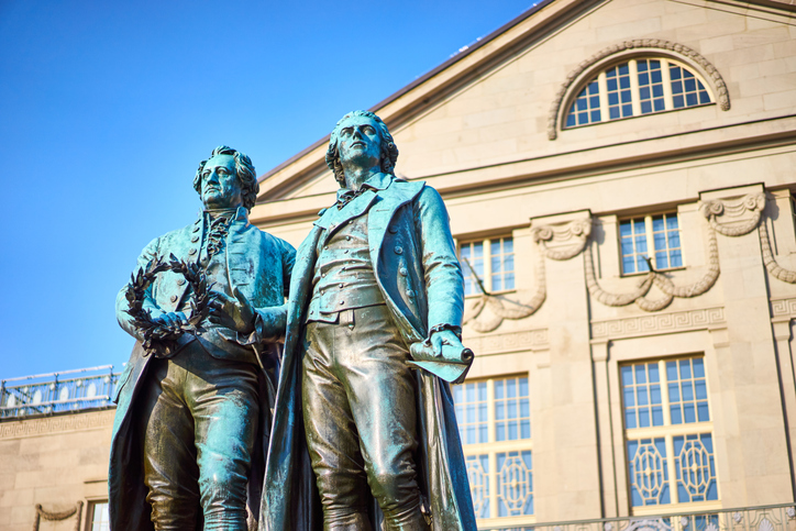 Das Goethe-und-Schiller-Denkmal steht vor einem klassizistischen Gebäude in Weimar unter blauem Himmel.