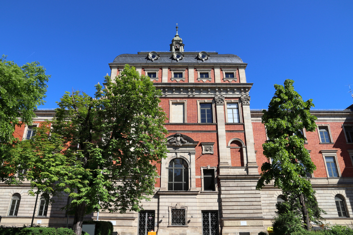 Repräsentatives historisches Gebäude in Erlangen mit roter Backsteinfassade, Natursteinelementen, großen Fenstern und grünen Bäumen vor blauem Himmel.