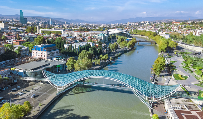 Luftaufnahme von Tiflis mit der modernen Friedensbrücke über den Kura-Fluss, grünen Uferbereichen und ausgedehnter Stadtlandschaft.