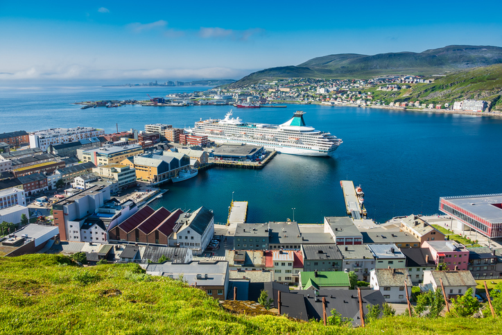 Großes Kreuzfahrtschiff im Hafen von Hammerfest mit Stadtgebäuden, ruhigem blauem Wasser und grünen Bergen im Hintergrund.