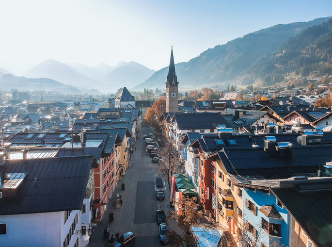 Blick über Kitzbühel mit Kirchturm, bunten Häusern und Bergen im Hintergrund bei leichtem Dunst.