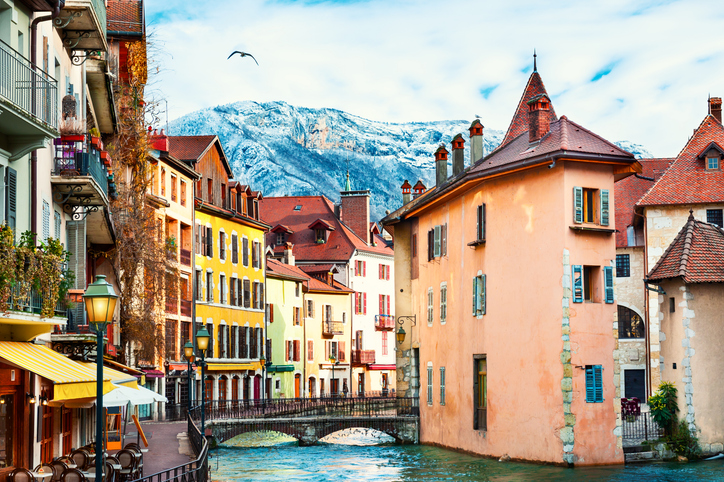 Bunte Häuser stehen an einem Kanal in Annecy, im Hintergrund erhebt sich ein schneebedeckter Berg.