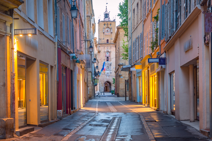 Eine schmale, ruhige Straße in Aix-en-Provence führt zwischen hohen Altstadthäusern auf einen historischen Turm zu.