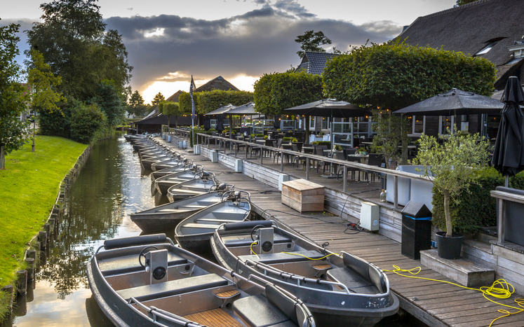 Mehrere kleine Boote liegen an einem Holzsteg vor einer Terrasse am Kanal in Giethoorn bei Abendstimmung.
