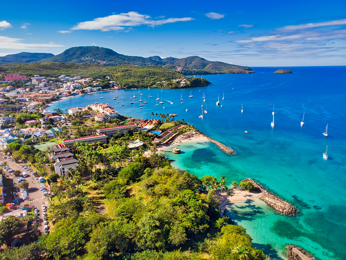 Luftaufnahme einer Bucht auf Martinique mit türkisblauem Wasser, Segelbooten, grünen Hügeln und Hotelanlage an der Küste.