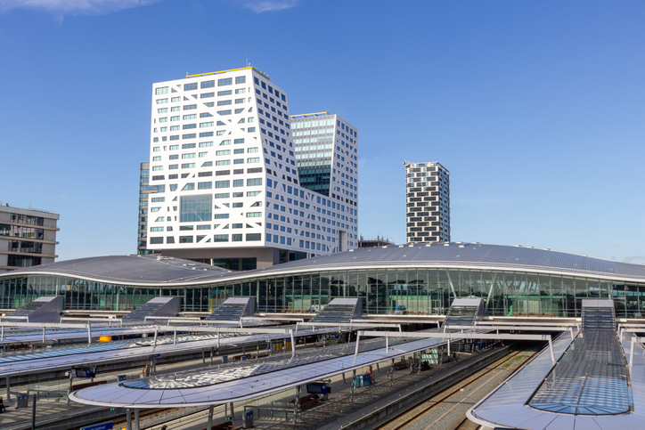 Utrecht Centraal mit modernen Bahnsteigen, großem Glasdach und hohen Bürogebäuden unter blauem Himmel.