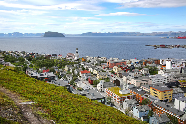 Blick über Hammerfest in Nordnorwegen mit bunten Häusern, Kirche, Hafen, Meer und karger Berglandschaft unter blauem Himmel.