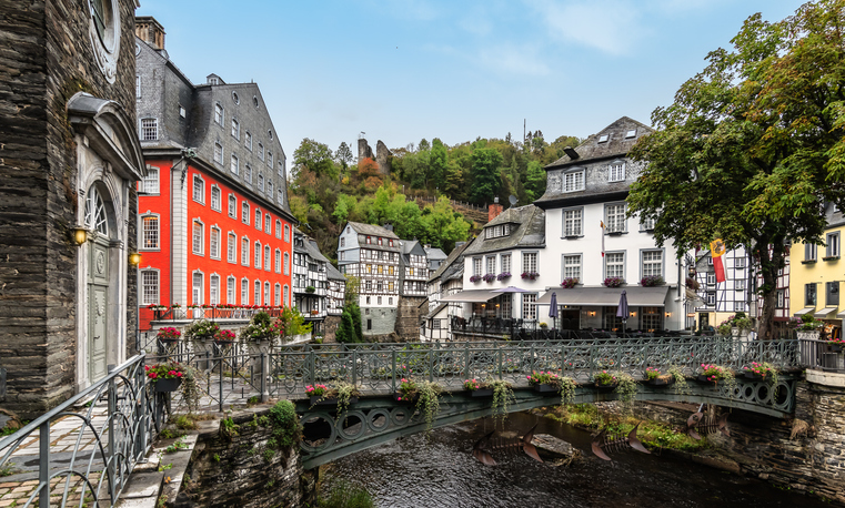 Kleine Brücke in Monschau über die Rur mit historischen Häusern, roter Fassade, Schieferdächern und begrüntem Hang im Hintergrund.