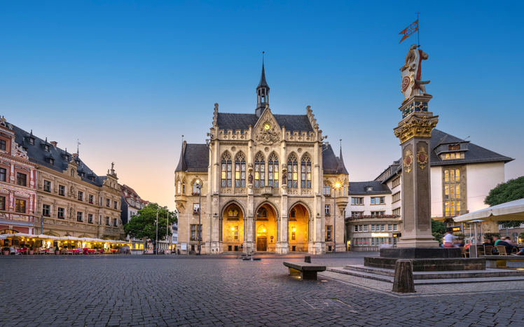 Historisches Rathaus am Fischmarkt in Erfurt mit beleuchteter Fassade, gepflastertem Platz und Säule im Vordergrund bei Abenddämmerung.