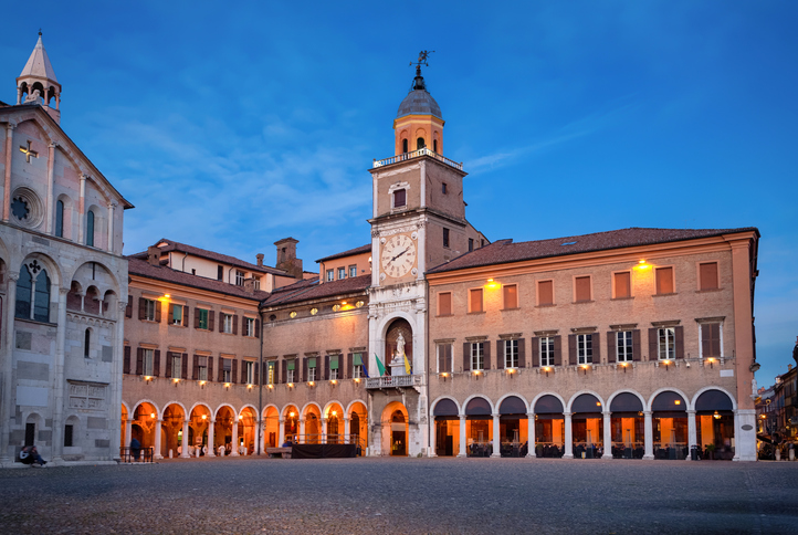 Piazza Grande in Modena am Abend mit historischem Palazzo Comunale, Uhrturm, beleuchteten Arkaden und gepflastertem Platz.