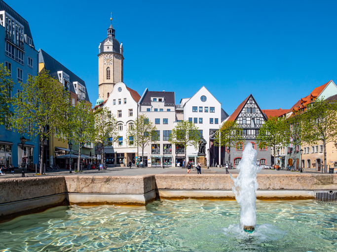 Ein Brunnen im Vordergrund, dahinter ein offener Platz mit historischen Häusern und einem markanten Turm in Weimar.