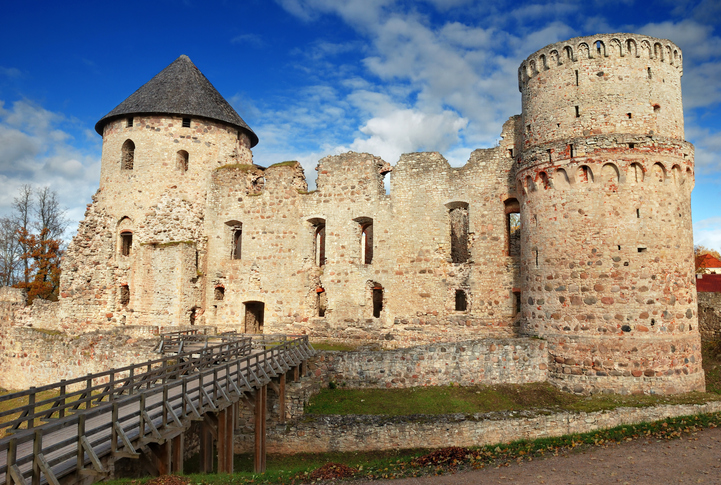 Historische Burgruine in Cēsis in Lettland mit massiven Rundtürmen, alten Steinmauern und hölzerner Brücke unter blauem Himmel.
