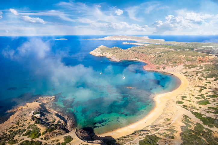 Luftaufnahme einer türkisblauen Bucht mit Sandstrand und felsiger Küste bei Ciutadella