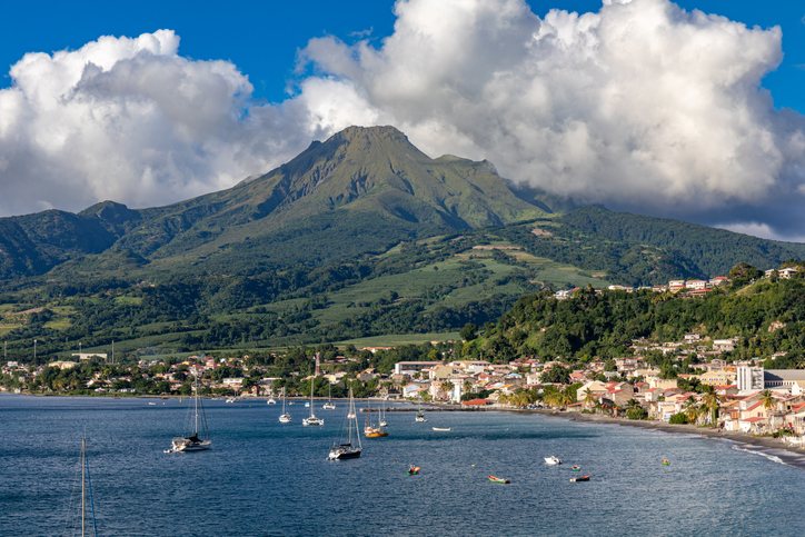 Blick auf die Küste von Martinique mit kleinen Booten im Wasser, einem Ort am Ufer und dem Vulkankegel des Mont Pelée im Hintergrund.
