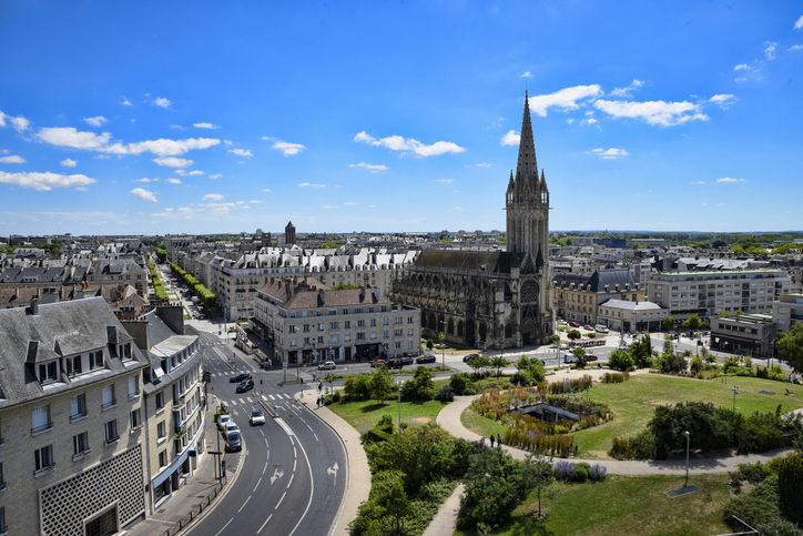 Blick über Caen mit großer gotischer Kirche, breiten Straßen, Grünflächen und moderner wie historischer Bebauung unter blauem Himmel.