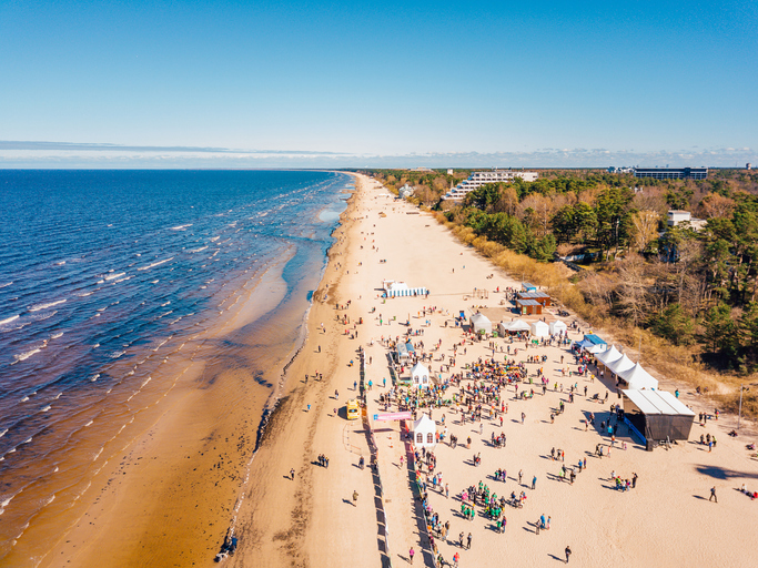 Luftaufnahme von Jūrmala mit langem Sandstrand, Ostsee, zahlreichen Besuchern sowie Zelten und Veranstaltungsflächen direkt an der Küste.