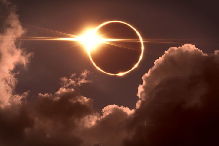 Fast vollständig vom Mond verdeckte Sonne mit hellem leuchtendem Ring und intensivem Licht zwischen dunklen Wolken.