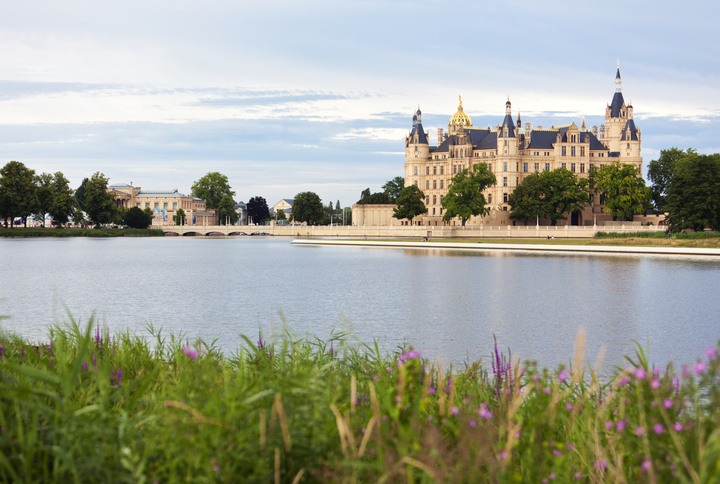 Das Schweriner Schloss steht prachtvoll am Ufer eines Sees, davor ziehen sich Wiesen und Blumen am Wasser entlang.