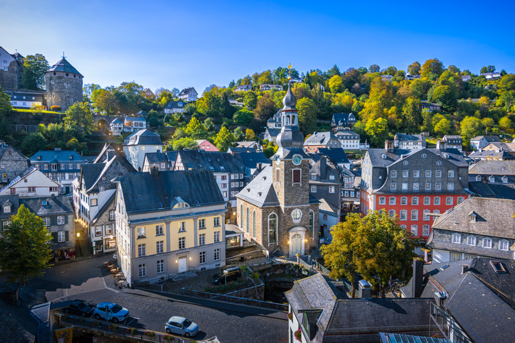 Blick über Monschau mit dichter historischer Bebauung, Kirche mit barocker Haube, Schieferdächern und bewaldeten Hügeln im Hintergrund.