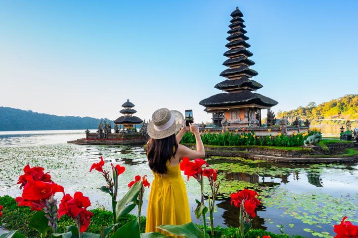 Frau in gelbem Kleid fotografiert einen balinesischen Tempel am See mit roten Blumen im Vordergrund
