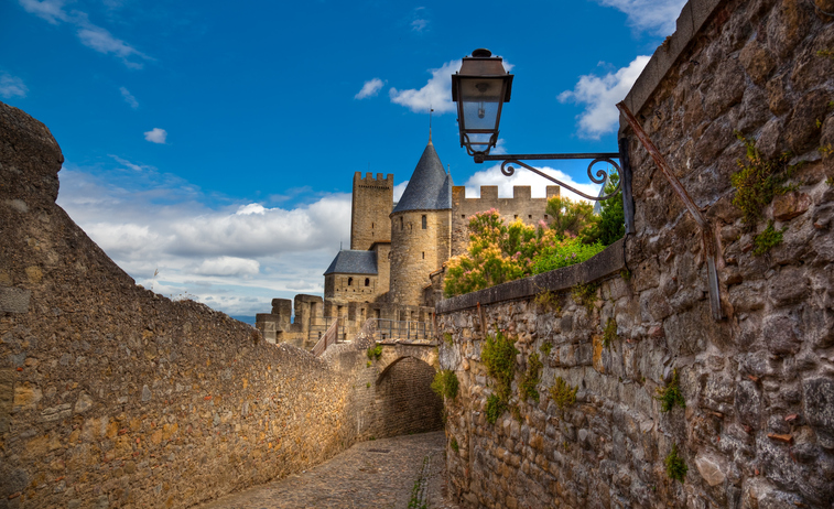 Ein schmaler gepflasterter Weg führt zwischen steinernen Mauern auf einen Turm der Festung Carcassonne zu.