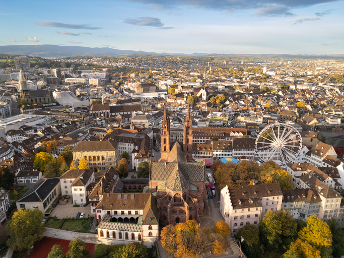 Luftaufnahme von Basel mit dem markanten Basler Münster, seinen beiden Türmen, historischen Altstadthäusern und einem Riesenrad im Stadtzentrum.