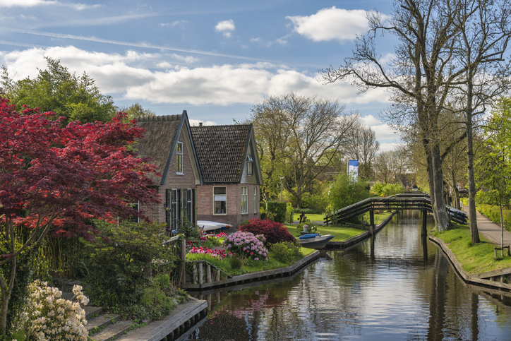 Ruhiger Kanal in Giethoorn mit Backsteinhaus, blühenden Sträuchern und kleiner schwarzer Brücke unter blauem Himmel.