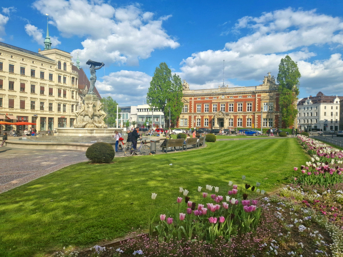 Gepflegte Grünanlage in Görlitz mit Blumenbeeten, Brunnen, Sitzbänken und historischen Gebäuden unter hellem Frühlingshimmel.