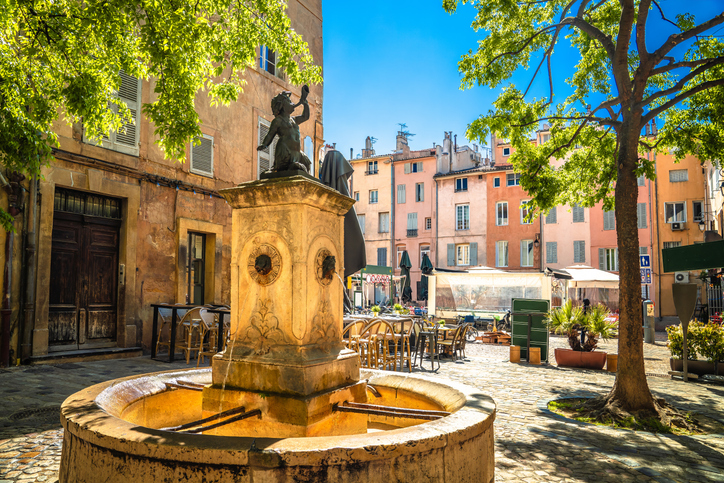 Ein kleiner steinerner Brunnen steht auf einem schattigen Platz in Aix-en-Provence, umgeben von Cafés und pastellfarbenen Häusern.