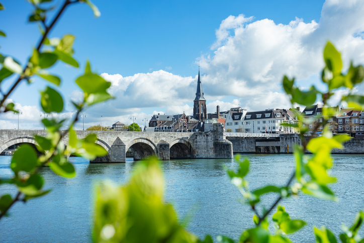 Steinerne Brücke über die Maas in Maastricht, eingerahmt von grünen Blättern, mit Stadtsilhouette und Kirchturm im Hintergrund.