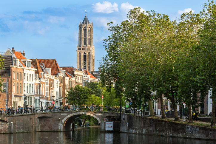 Gracht in Utrecht mit steinerner Brücke, historischen Häusern, Bäumen und dem Domturm im Hintergrund.