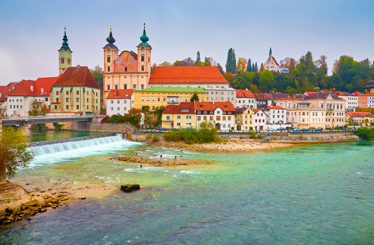 Panorama von Steyr mit Fluss, Wehr, Altstadthäusern und Kirchenanlage am Ufer.