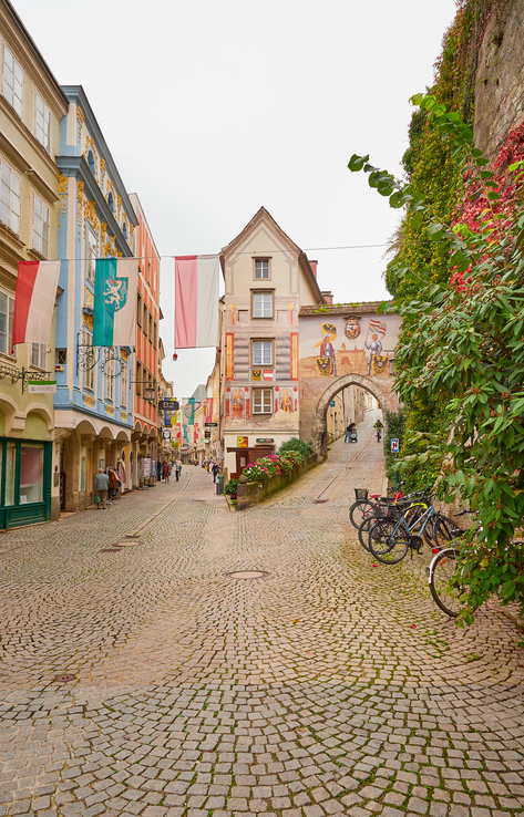 Kopfsteinpflasterstraße in Steyr mit bemalten Fassaden, Fahnen, Fahrrädern und einem Torbogen.