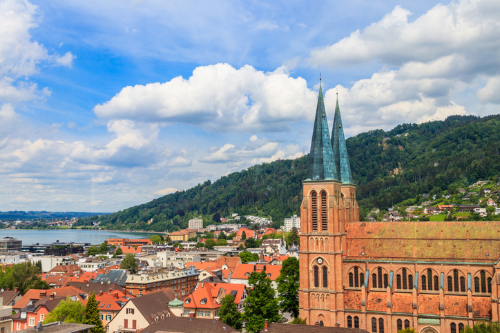 Panorama von Bregenz mit rotdachigen Häusern, Doppeltürmen der Pfarrkirche St. Gallus und bewaldetem Hang unter großem Wolkenhimmel.