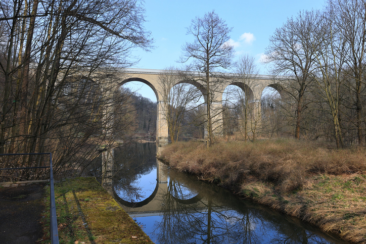 Hohes Viadukt bei Görlitz über einem ruhigen Flusslauf mit kahlen Bäumen und spiegelnder Wasseroberfläche an der Neißebrücke.