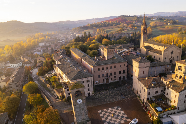 Historisches Zentrum von Castelvetro di Modena mit mittelalterlichen Gebäuden, Kirchturm, Schachbrettmuster auf der Piazza Roma und herbstlicher Hügellandschaft.