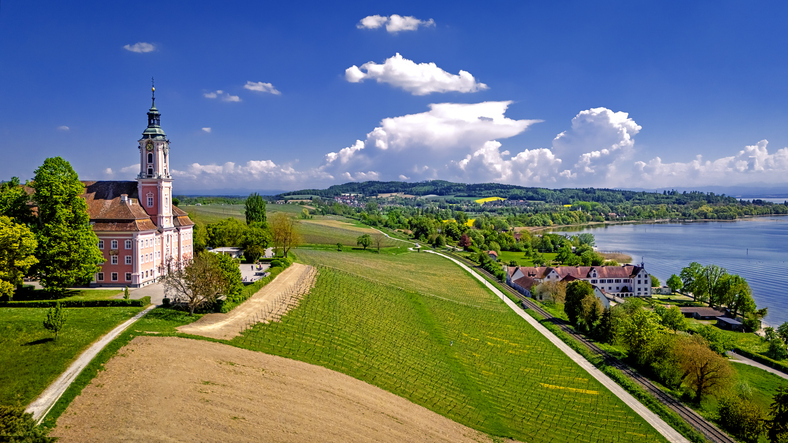 Eine barocke Kirche steht auf einer Anhöhe über Feldern und Weinbergen mit Blick auf den Bodensee.