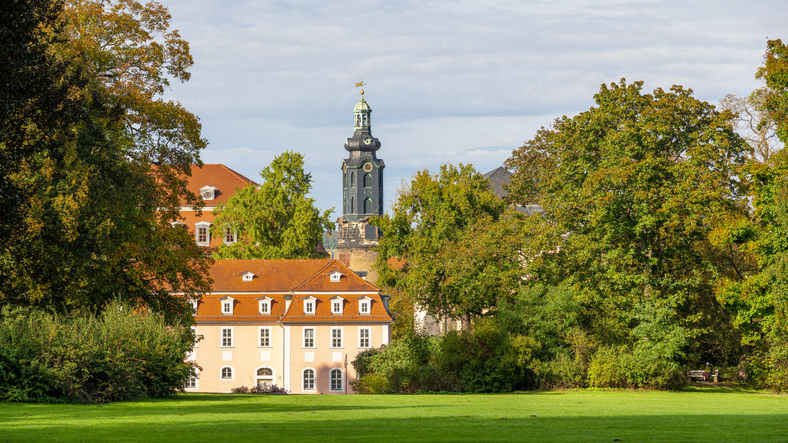 Große Wiese, historische Gebäude und ein dunkler Kirchturm prägen eine grüne Parklandschaft in Weimar.
