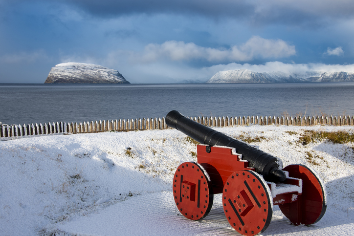 Historische schwarze Kanone mit roten Rädern an der verschneiten Küste von Hammerfest mit Meer, Inseln und schneebedeckten Bergen im Hintergrund.