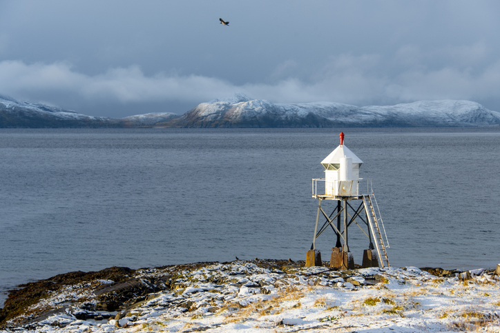 Kleines weißes Leuchtfeuer auf einer verschneiten felsigen Küste bei Hammerfest mit Meer und schneebedeckten Bergen im Hintergrund.