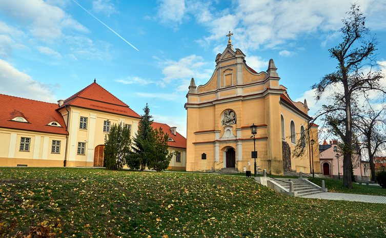 Historische Kirche in Gniezno mit heller Fassade, barockem Giebel, Nebengebäuden und Rasenfläche mit Herbstlaub.