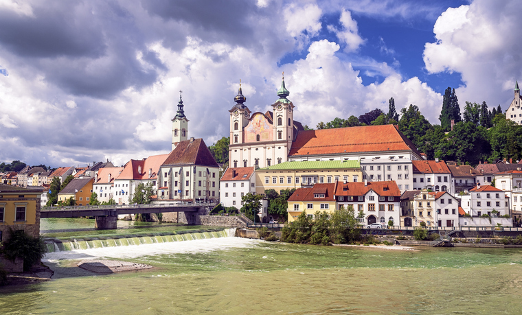 Historische Kloster- und Kirchengebäude über dem Fluss in Steyr unter dramatischem Wolkenhimmel.