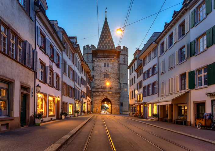 Historisches Spalentor in Basel zwischen Altstadthäusern mit Straßenbahnschienen und beleuchteten Geschäften in der Abenddämmerung.