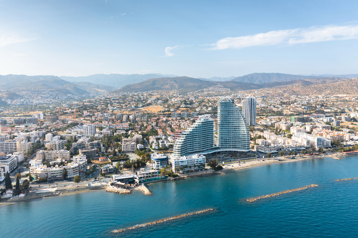 Luftaufnahme von Limassol mit moderner Hochhausarchitektur direkt am Mittelmeer, dichter Stadtbebauung und Bergen im Hintergrund.