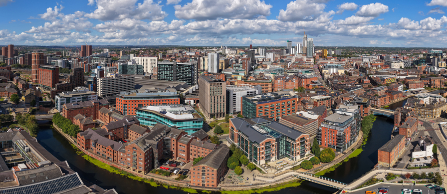 Luftaufnahme von Leeds mit River Aire, historischen Backsteingebäuden, modernen Wohnhäusern, Brücken und Hochhäusern im Hintergrund.