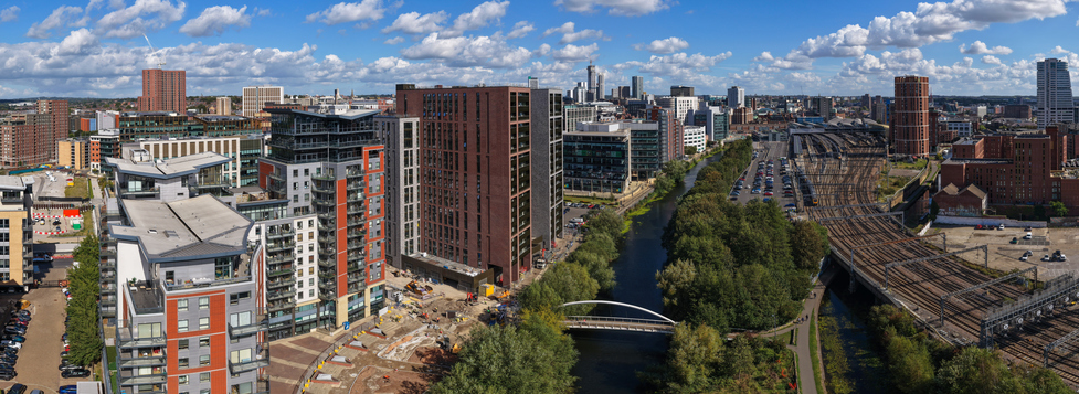 Panoramablick über Leeds mit modernen Wohn- und Bürogebäuden, Flusslauf, Brücke, Bahngleisen und Hochhäusern unter wolkigem Himmel.