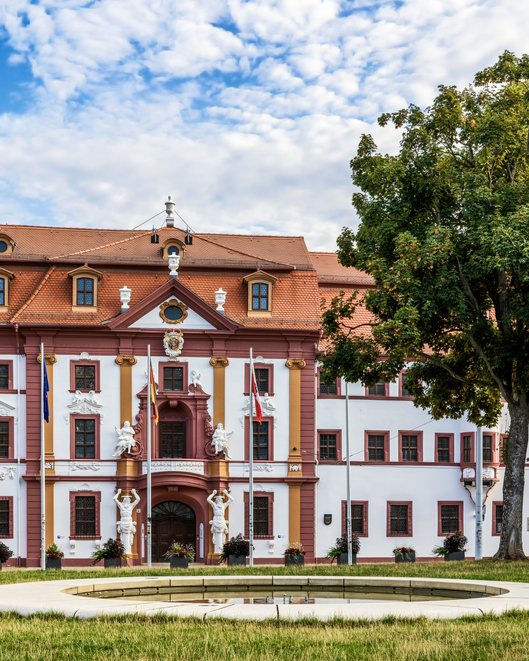Repräsentatives historisches Gebäude in Erfurt mit weiß-roter Fassade, barockem Eingangsbereich, Fahnen und gepflegter Grünfläche davor.