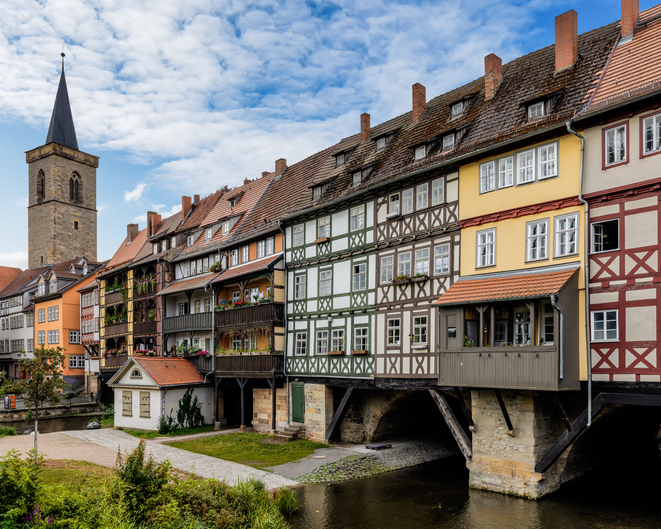 Fachwerkhäuser an der Krämerbrücke in Erfurt mit Flusslauf, kleinem Weg und Kirchturm im Hintergrund.
