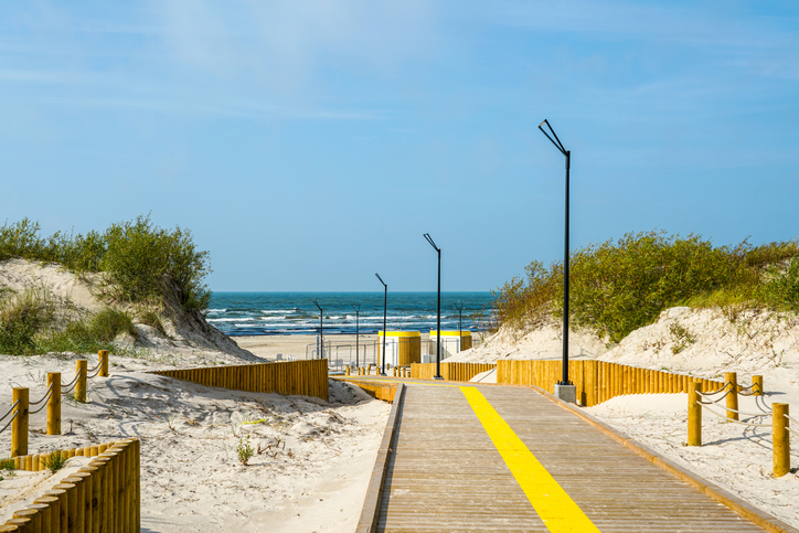 Moderner Holzsteg durch helle Sanddünen zum Ostseestrand von Liepāja in Lettland mit Meerblick und blauem Himmel.