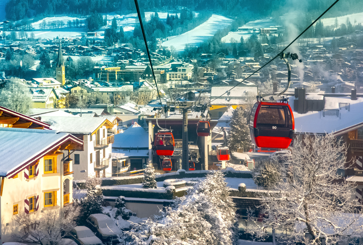 Rote Gondeln schweben über ein verschneites Kitzbühel mit Häusern, Bäumen und winterlicher Berglandschaft im Hintergrund.
